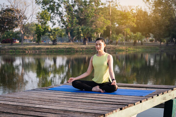Fototapeta na wymiar Young Asian beautiful woman practicing yoga and meditate in the lotus pose outdoor beside the lake in the morning for relaxation and peace of mind. Harmony and meditation concept. Healthy lifestyle