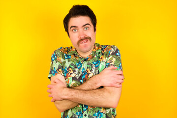 young handsome Caucasian man wearing Hawaiian shirt against yellow wall bitting his mouth and looking worried and scared crossing arms, worry and doubt.