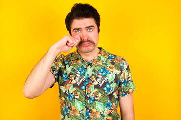 Unhappy young handsome Caucasian man wearing Hawaiian shirt against yellow wall crying while posing at camera whipping tears with hand.