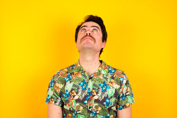 young handsome Caucasian man wearing Hawaiian shirt against yellow wall looking up as he sees...