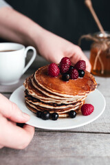 Hands hold white plate with pancakes, berries, honey, coffee cup on wooden table