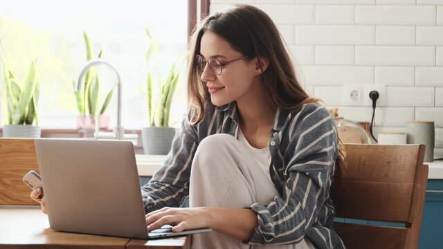A beautiful young woman wearing glasses is using her smartphone while working with her laptop computer sitting at the table in the kitchen at home