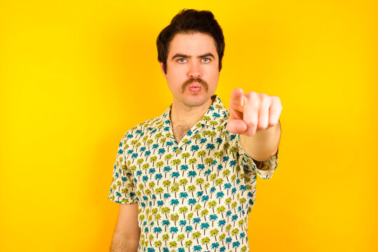 Cheerful young handsome Caucasian man wearing Hawaiian shirt against yellow wall indicates happily at you, chooses to compete, has positive expression, makes choice.