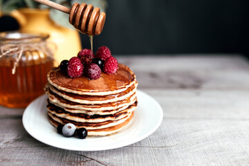 Juicy pancakes with berries and honey on a white plate, spoon, jar, wooden table