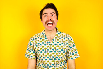 Surprised young handsome Caucasian man wearing Hawaiian shirt against yellow wall, shrugs shoulders, looking sideways, being happy and excited. Sudden reactions concept.