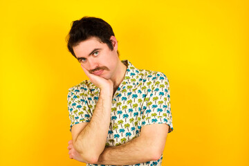 Very bored young handsome Caucasian man wearing Hawaiian shirt against yellow wall holding hand on cheek while support it with another crossed hand, looking tired and sick.