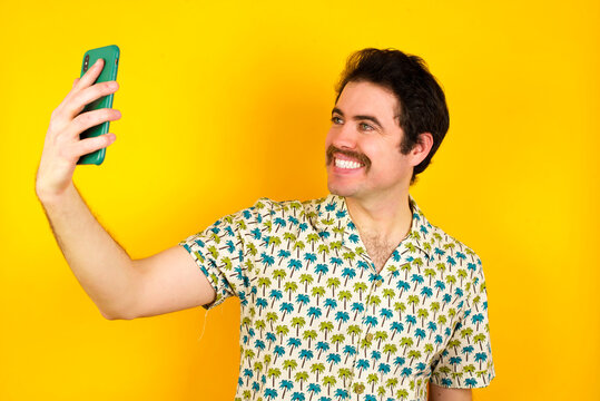 Portrait of a young handsome Caucasian man wearing Hawaiian shirt against yellow wall  taking a selfie to send it to friends and followers or post it on his social media.
