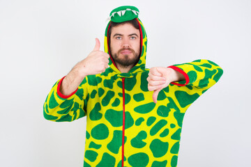 young caucasian man wearing a pyjama standing against white wall showing thumbs up and thumbs down,...