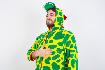 young caucasian man wearing a pyjama standing against white wall touches tummy, smiles gently,...