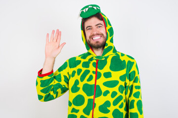 young caucasian man wearing a pyjama standing against white wall Waiving saying hello happy and smiling, friendly welcome gesture. © Jihan
