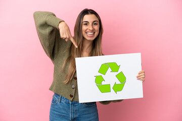 Young caucasian woman isolated on pink background holding a placard with recycle icon and  pointing...