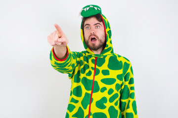 young caucasian man wearing a pajama standing against white wall Pointing with finger surprised ahead, open mouth amazed expression, something on the front.