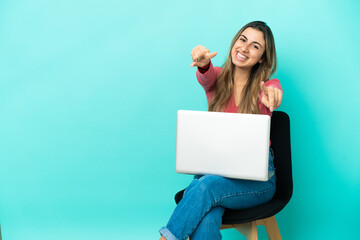 Young caucasian woman sitting on a chair with her pc isolated on blue background points finger at you while smiling