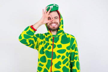 Fototapeta premium young caucasian man wearing a pajama standing against white wall doing ok gesture with hand smiling, eye looking through fingers with happy face.