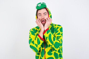 Fototapeta premium Stupefied young caucasian man wearing a pajama standing against white background expresses excitement and thrill, keeps jaw dropped, hands on cheeks, has eyes popped out