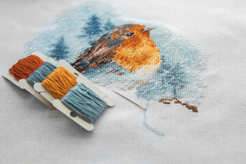 view of the cross stitch process multicolored bird, white canvas, floss threads on bobbins, needle,...