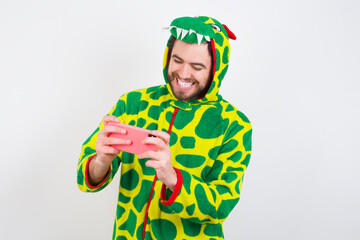 young caucasian man wearing a pajama standing against white background holding in hands cell playing video games or chatting