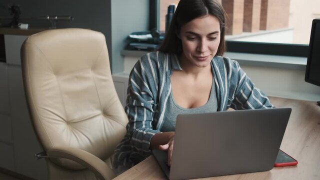 A pleased young woman is using her laptop computer while sitting in the office