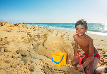 Portrait of a boy build sand castle on the sea beach on vacations sit with shovel and bucket