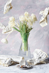 A bouquet of white tulips in a glass vase, spilled coffee and crumpled torn sheets with notes in the background and in the air.