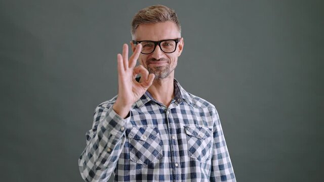 A pleased adult man wearing eyeglasses is showing ok gesture isolated over a grey background in the studio