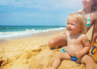 Face portrait of blond toddler boy sit in the sand happy smiling looking at the sea with hairs fly on from warm wind