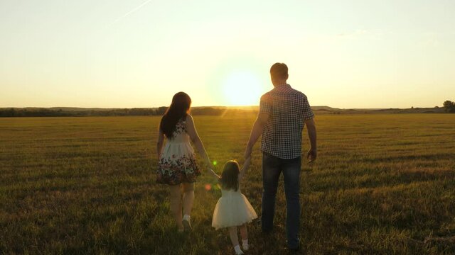 Happy family and little daughter play with mom and dad holding hands at sunset on field in park. Baby plays with his parents child is jumping in sun. Child and parent are walking in outdoors. Travel