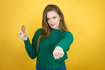 Young beautiful woman eating chocolate cookie over yellow background pointing with finger to the camera and to you, confident gesture looking serious