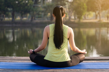 Young Asian beautiful woman practicing yoga and meditate in the lotus pose outdoor beside the lake in the morning for relaxation and peace of mind. Harmony and meditation concept. Healthy lifestyle