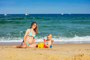 Toddler boy sit and pregnant young mother on the sand beach playing together near sea waves pointing with finger