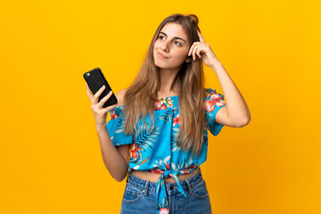 Young woman using mobile phone over isolated yellow background having doubts and with confuse face...