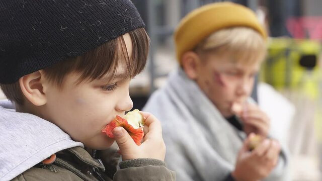 Homeless boys sit eating food on the bench, little caucasian children, street kids are hungry, get some food by kind people.