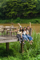 Three happy young Asian female friends sitting enjoying themselves on the wooden deck  by a river with big trees in background