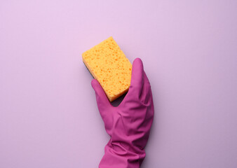 female hand in a pink rubber glove holds a yellow kitchen sponge on a purple background
