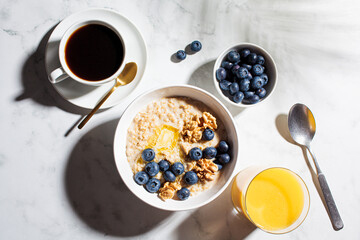 Oatmeal bowl with berries and nuts, cup of coffee and glass of orange juice on white marble background.