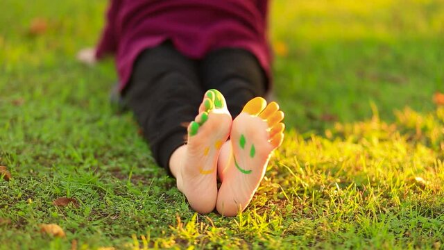 Little girl sits on grass and wounds legs which painted in funny smile faces. Summer time