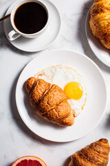 Breakfast food concept. Fried egg with croissant and cup of coffee on white marble background, vertical.