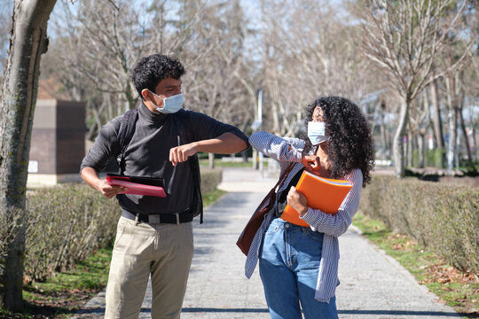 Two latin students wearing face mask and doing new social distancing greet with elbows bumps for preventing corona virus spread. New normal in the university campus.