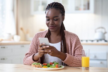 Smiling Black Woman Texting On Smartphone While Having Breakfast In Kitchen