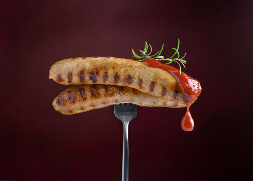 Grilled Sausages With Tomato Sauce And Rosemary On Fork On Dark Burgundy Background. Red Sauce Dripping From Sausages. Close-up.