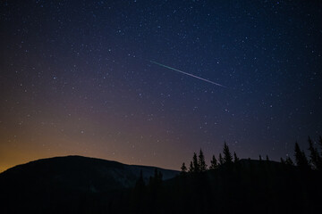 One meteor (shooting star) from the Perseids meteor shower on a summer night with a clear sky full of stars in the mountains with soft orange light from the city on the horizon, Kananaskis, Canada