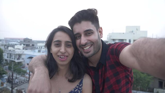 Smiling young Indian couple standing on rooftop and happily posing for selfie.Happy girl and cheerful guy having fun making selfie video on smart phone.Young smiling happy couple taking selfie. 