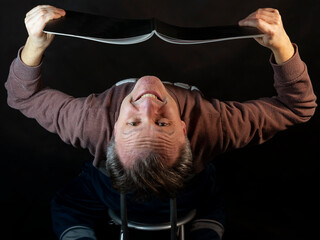 A middle-aged man reads a book with his head upside down with a witty expression
