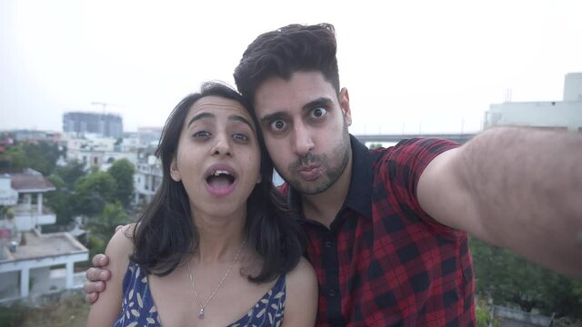 Joyful girl and guy taking funny selfie,having fun making funny faces.Cheerful and beautiful Indian couple taking selfie portrait on rooftop.Young funny couple taking selfie photos with smart phone
