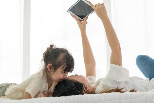 Asian Kid Kissing Her Mom While The Mother Using Tablet To Taking Photo Of Them On Bed. Family Together Concept