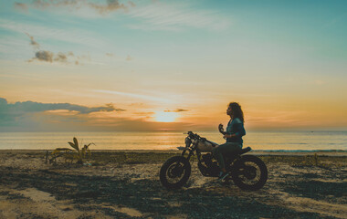 Beautiful girl having fun driving her custom cafe racer motorcycle, enjoying the sunset on the beach