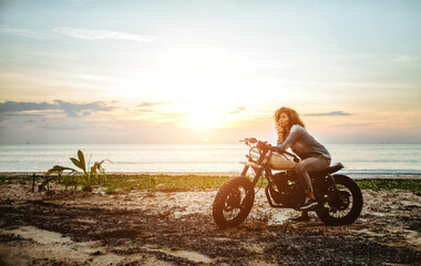 Mooi meisje dat plezier heeft met het besturen van haar aangepaste café racer-motorfiets, genietend van de zonsondergang op het strand