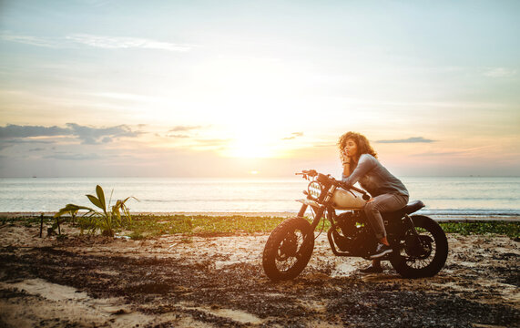 Beautiful girl having fun driving her custom cafe racer motorcycle, enjoying the sunset on the beach © oneinchpunch