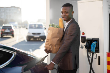 Portrait of young smiling black man in business clothes, holding paper shopping bag with food,...