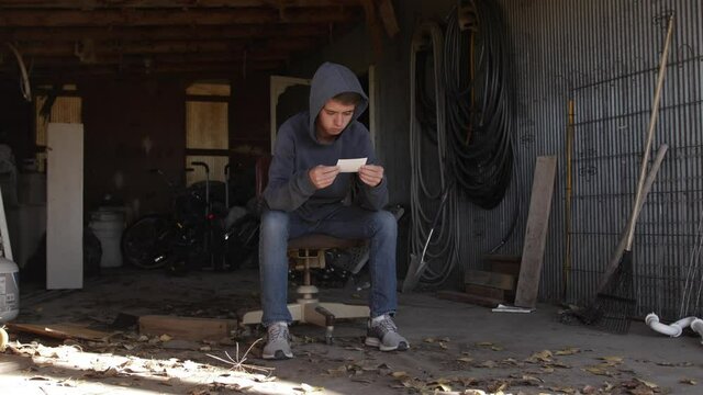 A teenage boy sits in a dirty garage looking at a picture. The young teen man looks sad and depressed and is wearing a hoodie.